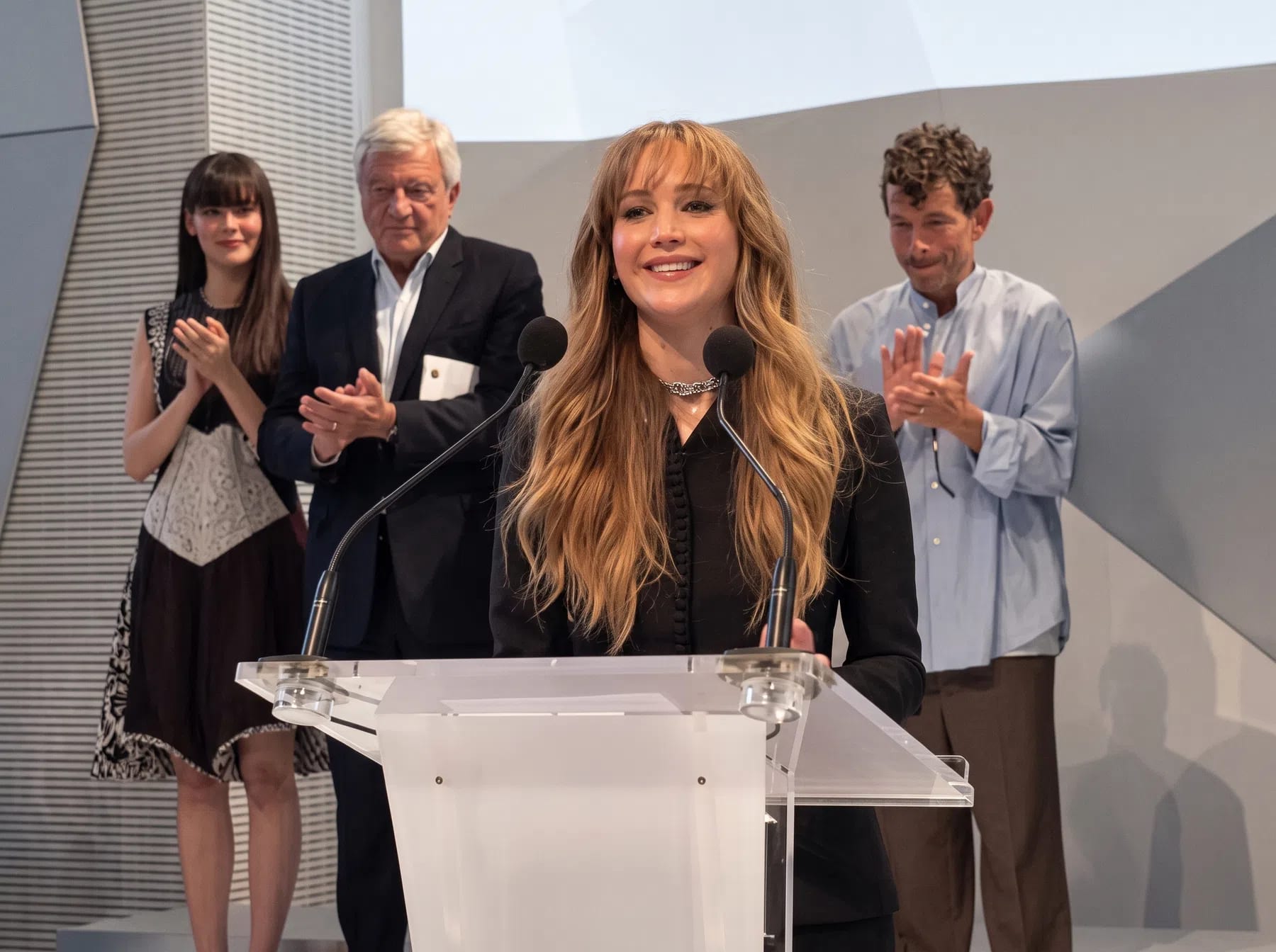 An eye-level medium shot captures American actress Jennifer Lawrence speaking smilingly from behind a transparent acrylic podium equipped with dual microphones. She has long wavy blonde hair with bangs and wears a tailored black jacket and a metallic choker. Standing in the background and applauding her are three individuals: a young woman on the left in a patterned dress, an older gentleman in a dark suit jacket, and a man on the right wearing a loose light blue button-down shirt and brown trousers. The setting features a modern, minimalist white stage with sharp geometric architectural angles.