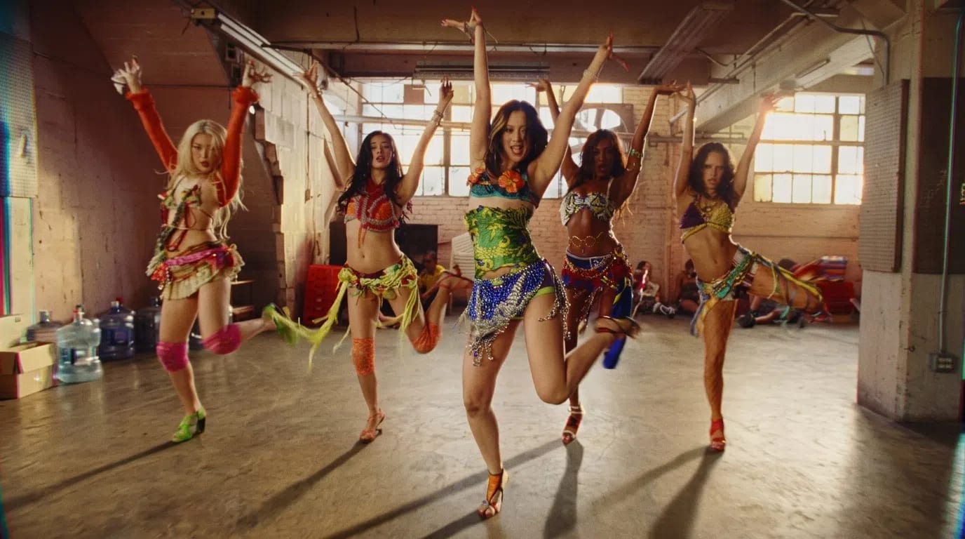 The global girl group KATSEYE performs high-energy, Latin-influenced choreography in a spacious, sunlit industrial warehouse for their vibrant music video "Hootie Frutti", appearing in mid-leap and dynamic dance poses while styled in heavily detailed, colorful Y2K-inspired festival attire featuring patterned crop tops, fringed wrap skirts, sashes, and mismatched leg and footwear accessories.
