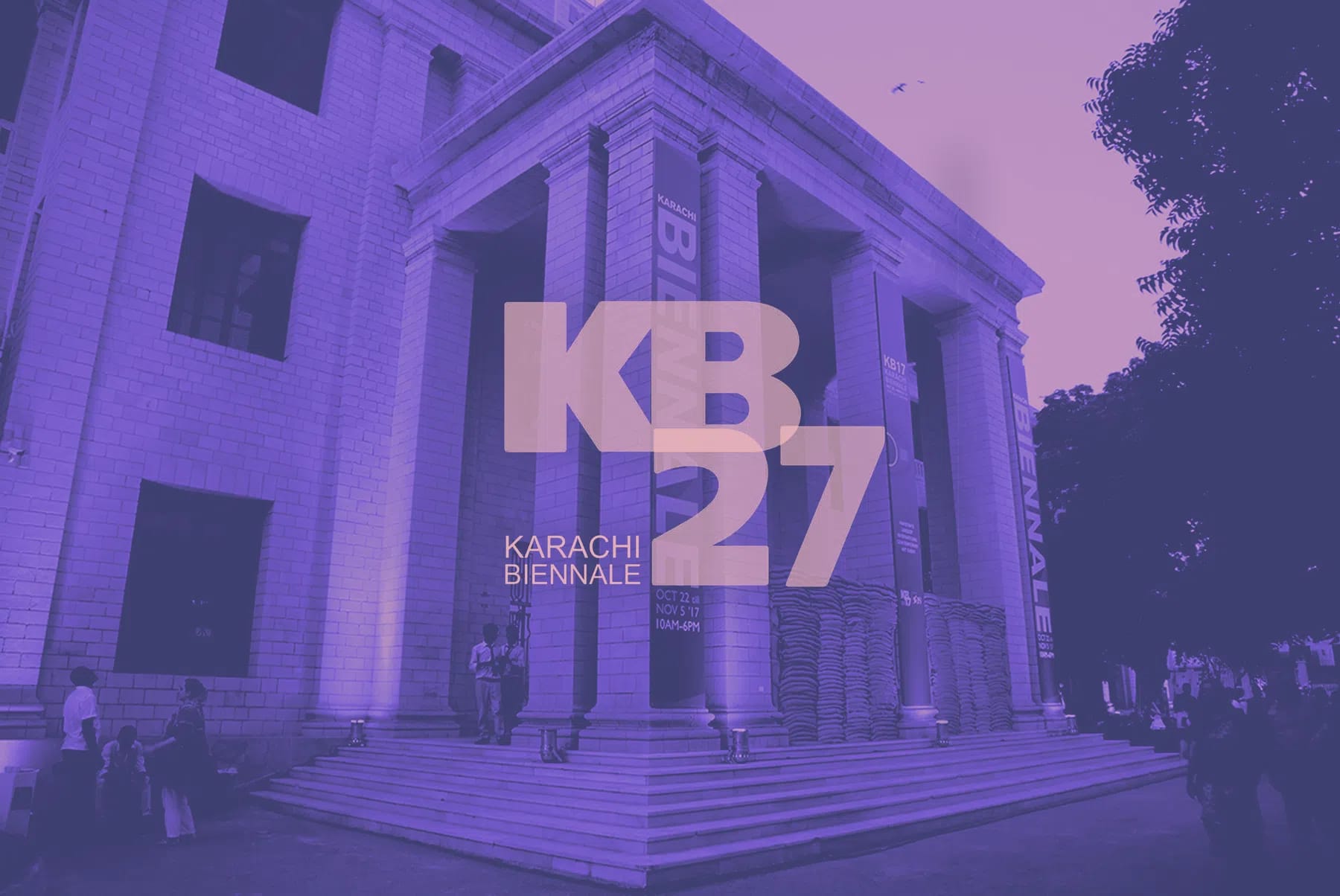 A large, grand building with towering stone pillars and wide concrete steps is bathed in a dramatic purple hue, serving as a backdrop for the Karachi Biennale. The architecture features tall rectangular windows and an expansive portico, with banners displaying "BIENNALE" hanging vertically down the columns. In front of the structure, a few individuals are scattered near the steps and base, appearing small against the monumental scale of the facade. Layered boldly over the center of the image is a large, stylized graphic text overlay reading "KB 27" and "KARACHI BIENNALE", accompanied by event dates indicating October 22 to November 5, '17, from 10 AM to 6 PM.