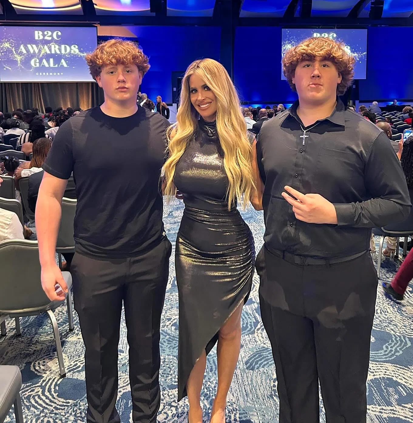 Kim Zolciak stands proudly between her two teenage sons, KJ and Kash Biermann, while attending the B2C Awards Gala Ceremony. The reality television personality wears a fitted, shimmering metallic dark grey gown featuring a mock neck and an asymmetrical thigh-high slit that highlights her figure. Her long blonde hair is styled in loose, voluminous waves draping over her shoulders. Flanking her on the left, one son wears a simple black crewneck t-shirt paired with matching black trousers, while the other son stands to her right in a black long-sleeved button-down shirt, a silver cross necklace, and matching black dress pants as he flashes a peace sign toward the camera. They pose together in a large, carpeted event hall filled with rows of seated attendees beneath bright blue overhead event lighting.