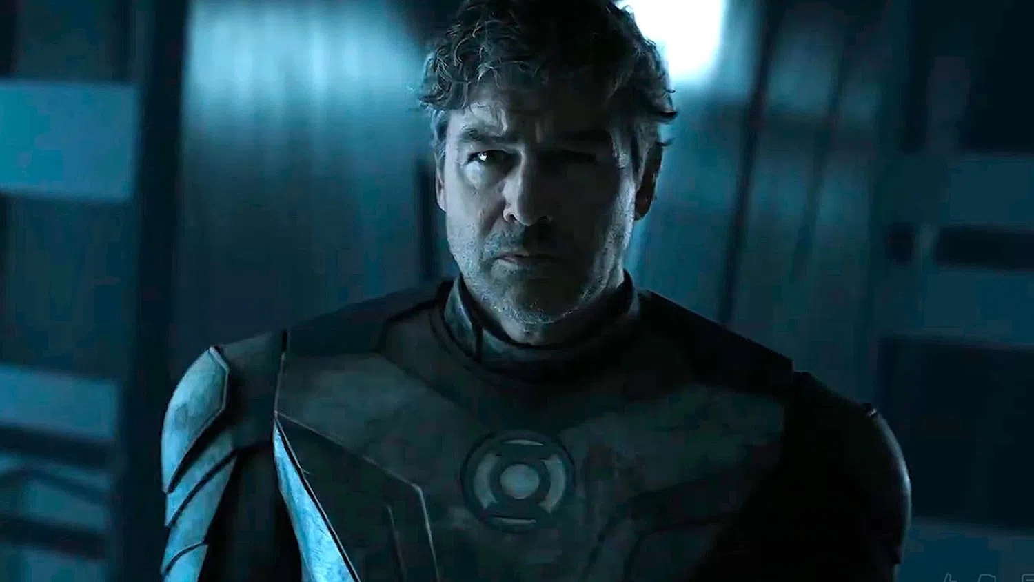Kyle Chandler stars as a veteran, grizzled Hal Jordan in the official promotional still from DC Studios’ highly anticipated HBO series, Lanterns. Bathed in cold, high-contrast blue and gray cinematic lighting, the seasoned intergalactic space cop sports messy, salt-and-pepper hair and light stubble, his stern expression reflecting the dark, grounded, detective-driven tone of the Earth-based murder mystery. His high-tech, tactical armored chest plate showcases a sleekly embedded, illuminated Green Lantern Corps logo right in the center. The heavily textured suit trades traditional bright green spandex for an oxidized, dark metallic aesthetic that matches the gritty, True Detective-inspired approach established by executive producers Chris Mundy, Damon Lindelof, and Tom King.
