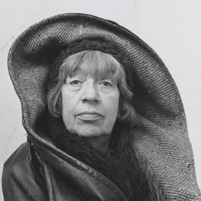 An archival, tight close-up, black-and-white portrait captures an elderly woman, identified as the influential American Abstract Expressionist painter Lee Krasner, looking directly at the camera with a steady, solemn expression. She features prominent, dark eyes under direct bangs, pronounced facial lines around her mouth, and full lips. Her head and shoulders are framed by an exaggerated, wide-brimmed straw or woven bonnet-style hat that dramatically curves upward and wraps around her head like a hood. Beneath the structured hat, a dark, textured knit or fur scarf is wrapped tightly around her neck, tucked into a dark leather or vinyl jacket that covers her shoulders. The high-contrast lighting accentuates the deep textures of her clothing, the woven patterns of the oversized headwear, and the weathered contours of her face against a minimalist, pale background.