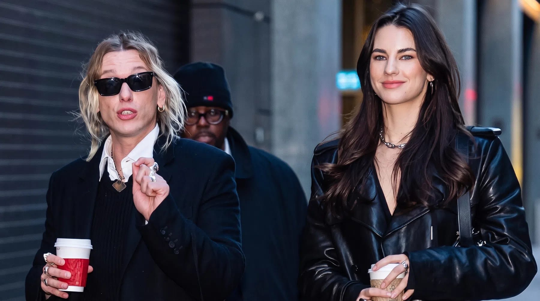 An eye-level, medium shot captures three individuals walking down a city street flanked by industrial buildings.On the left, a person with blonde, shoulder-length hair wears black sunglasses, a long black overcoat, a white collared shirt under a black sweater, and dark, pinstriped trousers. They hold a white paper coffee cup with a red sleeve in their right hand, capturing a candid, mid-speech expression.On the right, a woman with long, dark brown hair smiles forward while walking. She is dressed in a glossy black leather trench coat over dark gray trousers, holding a white coffee cup in her left hand.Slightly behind them and positioned in the center, a third individual wears a black beanie, glasses, and a dark coat, looking toward the left.The background features a gray, corrugated metal garage door on the left and a stone building facade on the right, casting soft, diffused daylight over the scene.