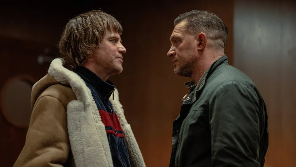 An intense promotional still from season 2 of the British crime drama series MobLand capturing actors Johnny Flynn and Tom Hardy standing face-to-face in a tense, profile confrontation against a dark wooden backdrop. On the left, Johnny Flynn (portraying Frankie) sports a shaggy, 1970s-style bowl-cut hairstyle and wears a heavy brown suede aviator jacket with a thick cream shearling fleece lining over a red, white, and navy blue track jacket. Facing him on the right, Tom Hardy (playing the underworld fixer Harry Da Souza) sports a tight, high-and-tight fade haircut and a rugged beard trim while wearing a dark green canvas utility jacket. Both characters exchange an unyielding, close-proximity gaze that underscores the escalating friction, power struggles, and impending civil war brewing between London's rival criminal factions.