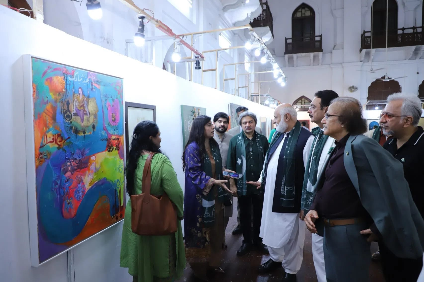 A vibrant indoor art exhibition features a diverse group of attendees gathered in a brightly lit, white-walled gallery space with high ceilings and traditional architectural balconies in the background. On the far left, a large, colorful, abstract painting filled with dense blue, orange, and green hues serves as a focal point. Several people, including a woman in a purple outfit and an older gentleman in a white shalwar kameez wearing a green scarf, stand in front of the artwork, actively engaged in a conversation while viewing the display. Other individuals, wearing formal attire or blazers, observe the gallery pieces under an overhead string of spotlights.