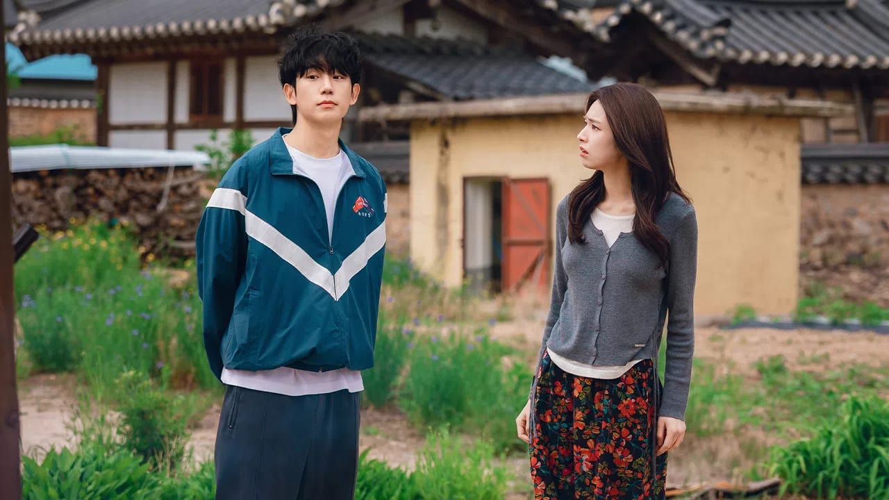 The image features South Korean actors Jung Hae-in and Ha Young starring in the Netflix romantic-comedy series Our Sticky Love. A young man and a young woman stand outdoors in a rural or historic village setting with traditional Korean-style tiled roofs visible in the background. The man on the left has messy black hair and wears a dark teal track jacket with a white chevron stripe across the chest over a white t-shirt, paired with dark grey sweatpants. He stands with his hands behind his back and a neutral, slightly distant expression, looking directly forward. To his right, the woman with long, dark brown hair stands in profile, looking at him with a slightly furrowed brow and a concerned or questioning facial expression. She wears a long-sleeved grey cardigan unbuttoned at the hem, a light-colored top underneath, and a vibrant, dark-toned skirt patterned with red and orange flowers. The background features a rustic dirt yard with sparse green patches, a weathered outbuilding with an open wooden door, and stone-accented structures that give the scene a cinematic, dramatic, or storytelling atmosphere.