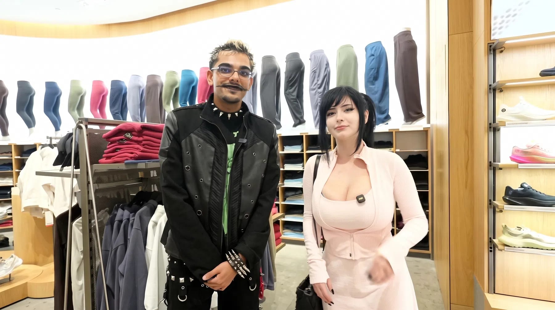 An overhead shot captures social media personalities Pinkchyu and N3on standing side-by-side inside a brightly lit Lululemon retail store, surrounded by displays of athletic apparel and footwear. On the left, N3on stands with his hands clasped forward, sporting a black jacket over a green graphic shirt, edgy spiked jewelry, and a thin, stylized mustache. To his right, Pinkchyu smiles toward the camera, dressed in a form-fitting, pale pink athletic zip-up jacket and matching top, with her dark hair styled in high pigtails. The background features neat rows of colorful leggings displayed on mannequins, stacks of folded hoodies, and a wooden shelf on the right showcasing various running shoes.