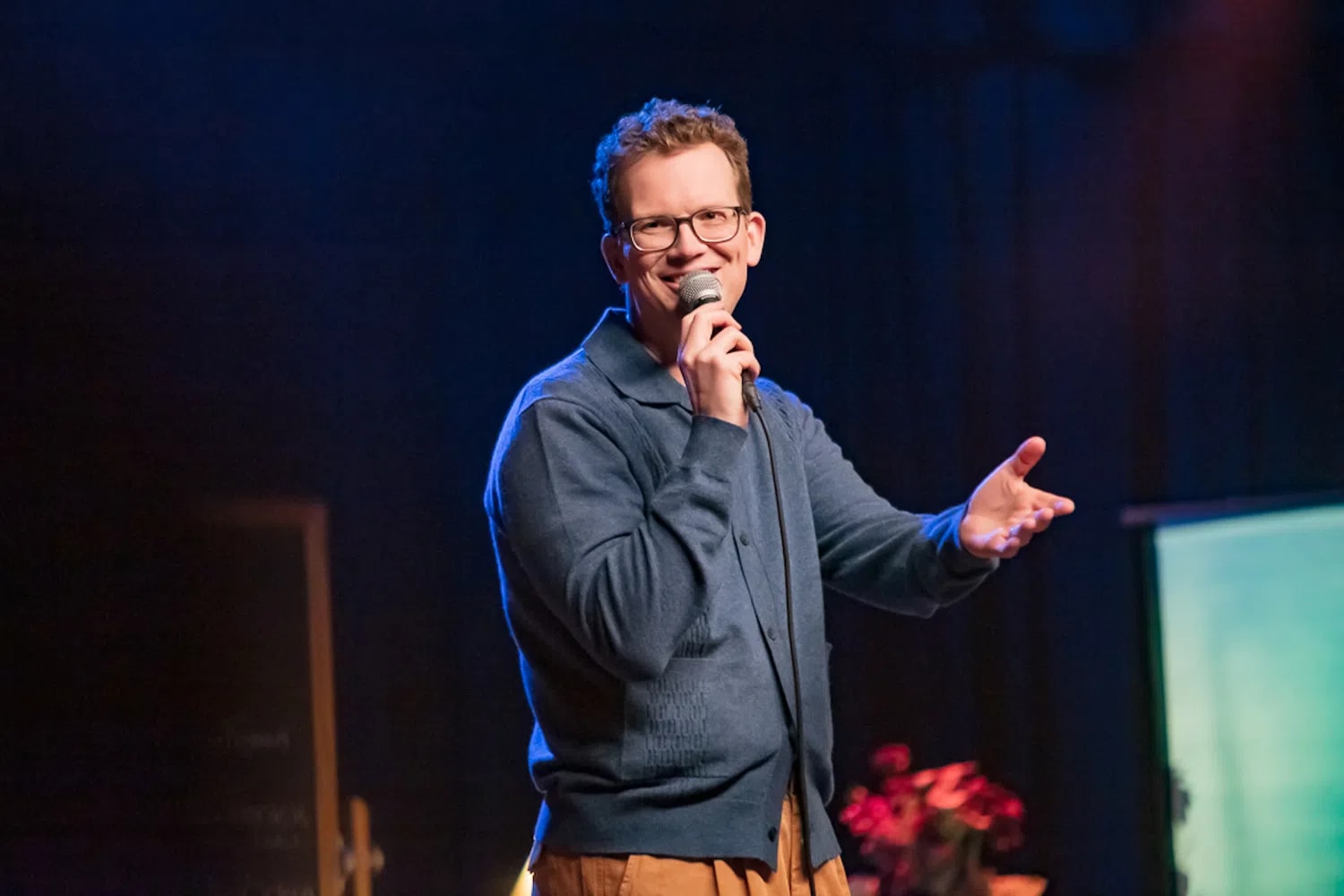 Popular internet educator and author Hank Green stands mid-gesture under vibrant stage lighting during a performance of his debut stand-up comedy special, Pissing Out Cancer, which premiered via Dropout Presents. Wearing his signature thick-rimmed glasses, a structured slate-blue buttoned cardigan polo, and casual tan trousers, he smiles broadly while holding a corded microphone close to his mouth with his right hand and extending his left hand outward to engage with the audience.