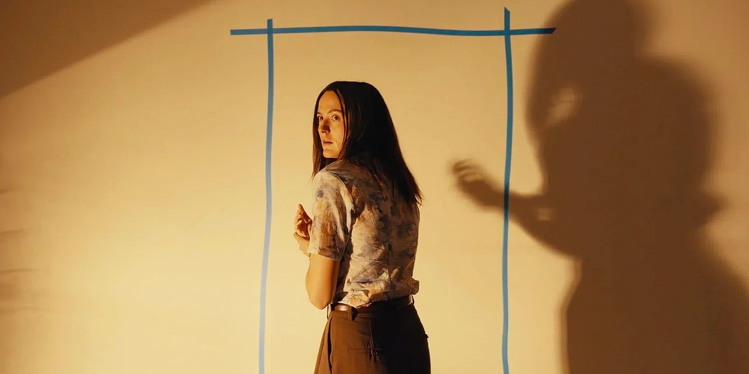 Renate Reinsve stars as Dr. Mary Kline in a suspenseful, dimly lit sequence from A24's psychological sci-fi horror feature Backrooms, looking back over her shoulder with an expression of intense vulnerability and fear. Clad in a patterned, short-sleeved short button-up shirt and high-waisted brown trousers, she stands frozen against a warm, yellow-toned wall bisected by a crude doorway outlined in bold blue tape. Looming aggressively over the right side of the frame is a stark, distorted human shadow cast onto the wall surface, its hand raised in a tense or demanding gesture. The sharp cinematic lighting emphasizes the depth of the shadow and highlights the actress's panicked profile, capturing the exact moment the film's domestic reality fractures into a surreal, multi-dimensional nightmare.