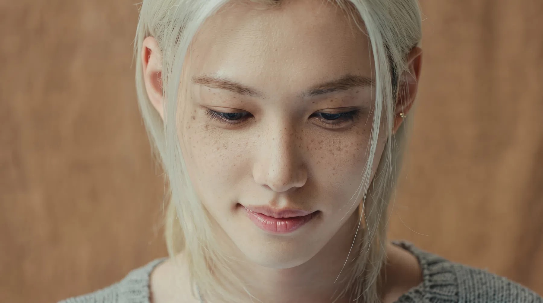 The image shows a close-up portrait of a young individual with striking features, captured from a slightly elevated angle as they look downward with a soft, serene expression. Their long, straight, platinum-blonde hair is parted down the middle, framing a face characterized by pale skin, dark, well-defined eyebrows, and prominent, natural freckles dusting the cheeks and bridge of the nose. Their eyes are cast down, and a gentle, subtle smile plays on their pink lips, giving the portrait a contemplative and calm demeanor. The lighting is soft and even, highlighting the smooth texture of the skin and the individual strands of hair, while the background is a muted, warm beige tone that keeps the focus entirely on the subject. In the lower-left corner, the vibrant blue "JYP" logo and white "OFFICIAL" text are visible, with the words "Slow It Down" written in a clean white serif font just above them.