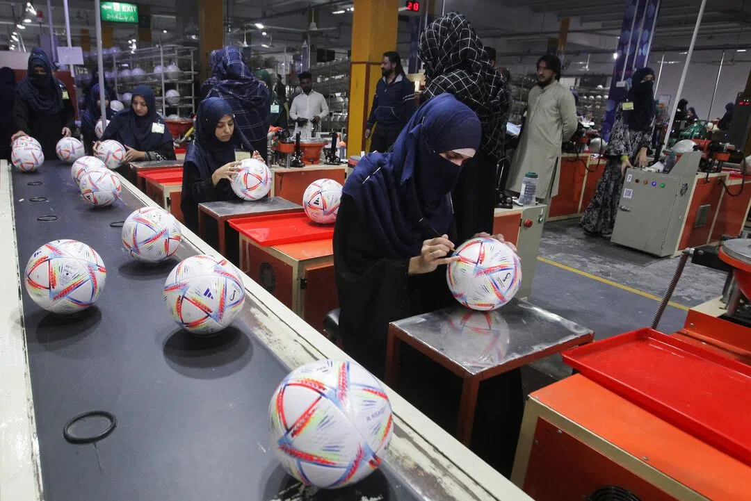 A wide shot shows the interior of a brightly lit manufacturing facility where workers, mostly women wearing hijabs and abayas, are meticulously assembling and inspecting official FIFA World Cup soccer balls on a production line. In the foreground, several white soccer balls with vibrant multi-colored geometric patterns travel along a gray conveyor belt, while a worker in a dark blue niqab uses a tool to fine-tune a ball resting on a metal stand. In the background, other workers are stationed at assembly tables under industrial ceiling fixtures, surrounded by machinery, equipment, and an exit sign visible in the distance.