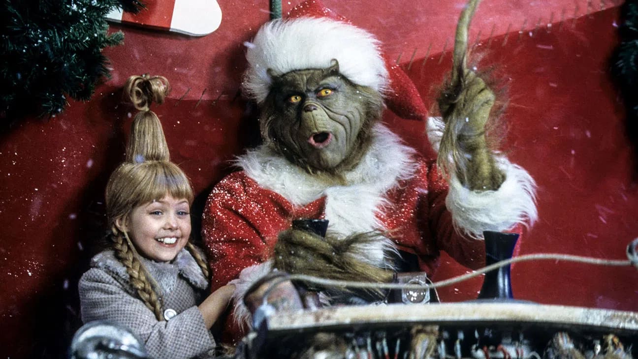 An image from the 2000 live-action film How the Grinch Stole Christmas features the Grinch, played by Jim Carrey in full green prosthetics and a Santa Claus suit, sitting in a makeshift sleigh next to a smiling, young Cindy Lou Who, played by Taylor Momsen, who sports her signature tall, braided updo and a grey winter coat amid a backdrop of falling snow and festive holiday decorations.