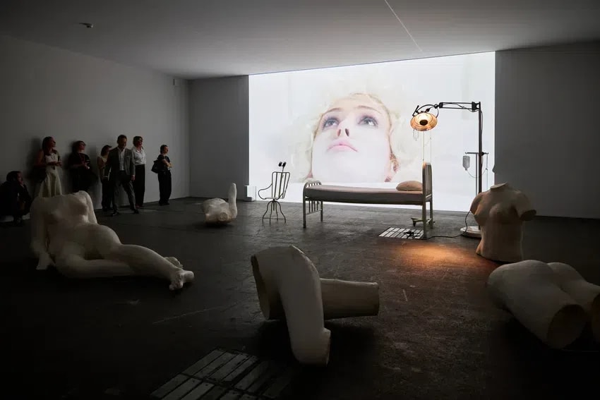 The image captures a haunting and cinematic art installation by Italian artist Vanessa Beecroft, characteristic of her exploration of the female form and the clinical gaze. The dimly lit gallery space is dominated by a large-scale video projection of a woman’s face, pale and ethereal, appearing as if in a state of suspended animation. On the floor and around the room, various disembodied, life-sized white sculptures—resembling fragmented mannequins or plaster casts—are scattered in poses of repose or abandonment. A stark, metal medical bed stands centrally before the projection, illuminated by a vintage operating theater lamp, which casts a warm, focused glow that contrasts with the cold, atmospheric lighting of the rest of the room. A small group of spectators stands along the left wall, their dark silhouettes observing the intersection of performance, sculpture, and video art.