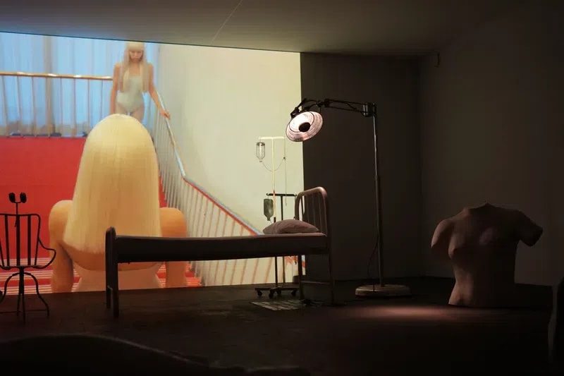 The image captures a haunting and cinematic art installation by Italian artist Vanessa Beecroft, characteristic of her exploration of the female form and the clinical gaze. The dimly lit gallery space is dominated by a large-scale video projection of a woman’s face, pale and ethereal, appearing as if in a state of suspended animation. On the floor and around the room, various disembodied, life-sized white sculptures—resembling fragmented mannequins or plaster casts—are scattered in poses of repose or abandonment. A stark, metal medical bed stands centrally before the projection, illuminated by a vintage operating theater lamp, which casts a warm, focused glow that contrasts with the cold, atmospheric lighting of the rest of the room. A small group of spectators stands along the left wall, their dark silhouettes observing the intersection of performance, sculpture, and video art.