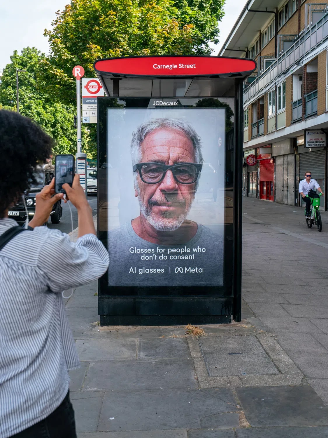 The image captures a street scene focused on a controversial mock advertisement displayed on a digital bus stop shelter or billboard. In the foreground on the left, a person with dark, curly afro hair, wearing a striped shirt, holds up a smartphone to take a picture of the ad. The advertisement features a large, close-up, grayscale portrait of convicted sex offender Jeffrey Epstein wearing a pair of thick, dark-rimmed smart glasses. Text printed in white at the bottom of the ad reads, "Glasses for people who don't do consent" followed by "AI glasses | ♾️ Meta", parodying Meta's Ray-Ban smart glasses marketing campaign. The ad is reflected in its glass casing, showing hints of the outdoor environment. The background reveals an urban sidewalk, a closed storefront with a red security shutter, and vehicles parked on the street under daylight.