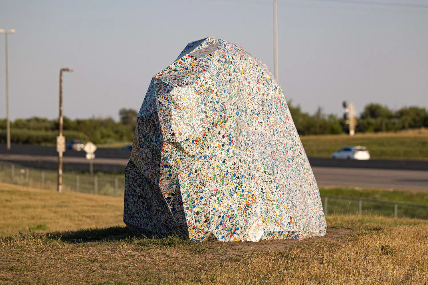 The image features "Modern Erratics," a controversial $297,490 permanent public art installation commissioned by the City of Saskatoon consisting of three large boulder-shaped sculptures crafted out of thousands of kilograms of compressed recycled plastic waste. Designed by the Calgary-based artist duo Sans façon to mimic glacial erratic boulders while making a statement on environmental pollution, this specific piece located in Diefenbaker Park is constructed from multi-colored recycled plastic bottle caps. The installation has sparked a heated public debate across Saskatchewan over tax spending, with some residents and the Canadian Taxpayers Federation mocking it as a waste of funds while city officials and arts advocates defend the project for promoting environmental awareness and community conversation.