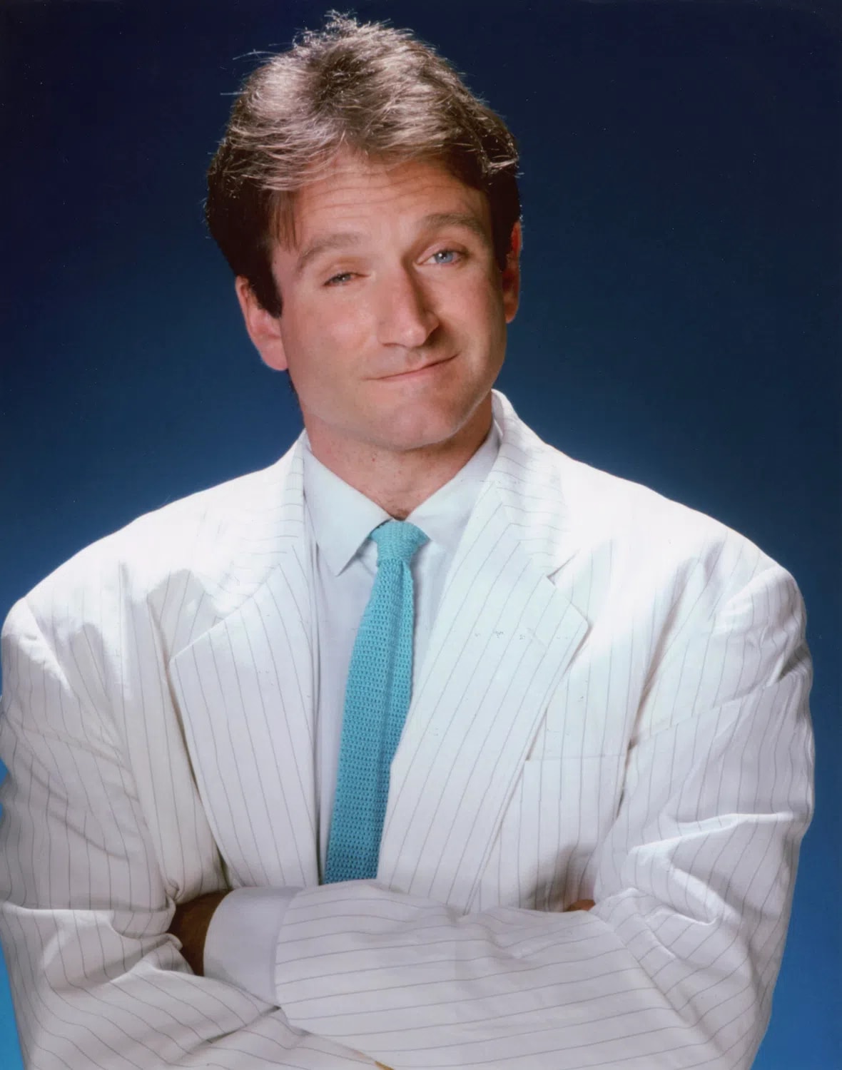 The image features a studio portrait of a young Robin Williams, captured from the chest up against a solid blue background. He is looking directly at the camera with a subtle, knowing smirk and slightly raised eyebrows, conveying his signature playful and charismatic energy. He is dressed in a stylish 1980s-style oversized white blazer with fine, light pinstripes, worn over a crisp white button-down shirt. A textured, light turquoise knit tie serves as a bright pop of color in the center of the frame. His arms are tightly crossed over his chest, and his dark brown hair is styled in a voluminous, feathered look characteristic of the era. The lighting is soft and even, highlighting his expressive facial features.