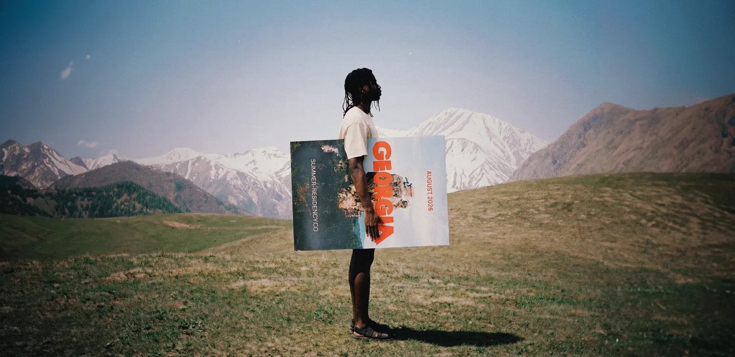 The image presents a wide, cinematic shot of a person standing sideways in a grassy field set against a backdrop of rolling green hills and large, snow-dusted mountains under a clear blue sky. The individual has long dark hair styled in braids or dreadlocks and wears a plain white shirt, dark shorts, and sandals. They are holding a large, rectangular poster or vinyl sleeve horizontally across their torso; the right side of the poster features the word "GEORGIA" written in large, bold red capital letters stacked vertically over a portrait of a face.