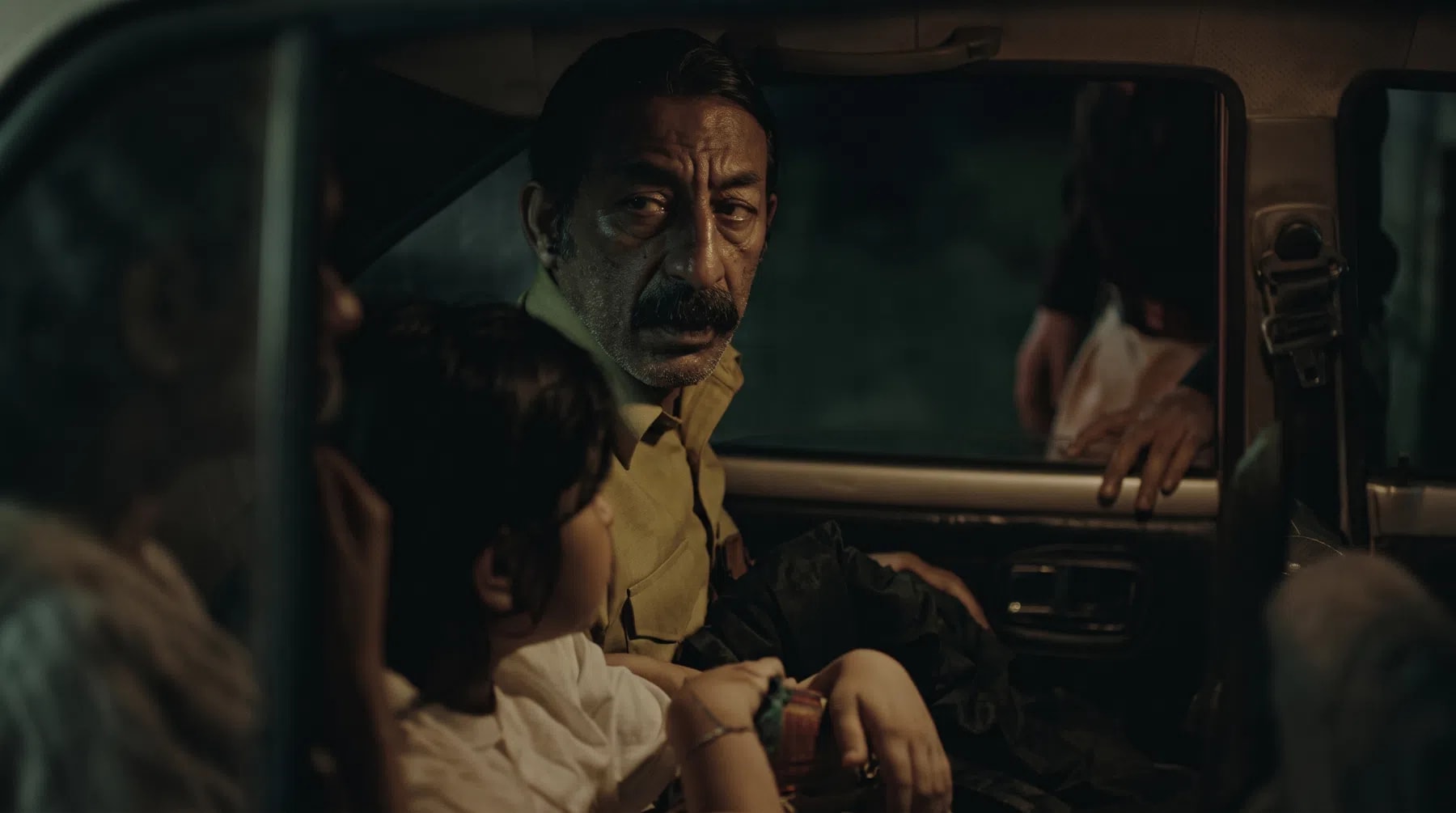 A dimly lit, cinematic film still captured from inside a vintage automobile at night showcases a tense and suspenseful sequence. The focal point of the composition is a middle-aged South Asian man with a prominent mustache and an anxious expression, dressed in a light-colored button-up shirt, looking warily out the open driver-side window. A hand rests heavily on the car's window frame from the outside, belonging to an unseen figure standing in the darkness, hinting at an active interrogation or confrontation. In the foreground, the back of a young child's head with dark hair is visible, seated next to another partially obscured passenger on the left, capturing a quiet, stressful moment shared within the vehicle's cramped interior.