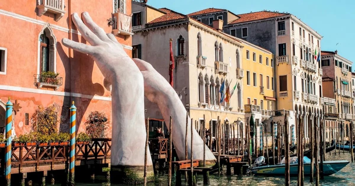 The monumental sculpture shown is "Support" (2017) by contemporary Italian artist Lorenzo Quinn. This detailed photograph captures a large-scale public art installation in Venice, featuring two massive, white, child-like hands emerging from the deep green waters of the Grand Canal. The hands are positioned as if they are literally supporting and bracing the historic, salmon-colored facade of the Ca' Sagredo Hotel. One hand grips the corner of the ornate building while the other reaches up against its side, set against a backdrop of sunlit Venetian architecture and traditional wooden mooring poles lining the canal.