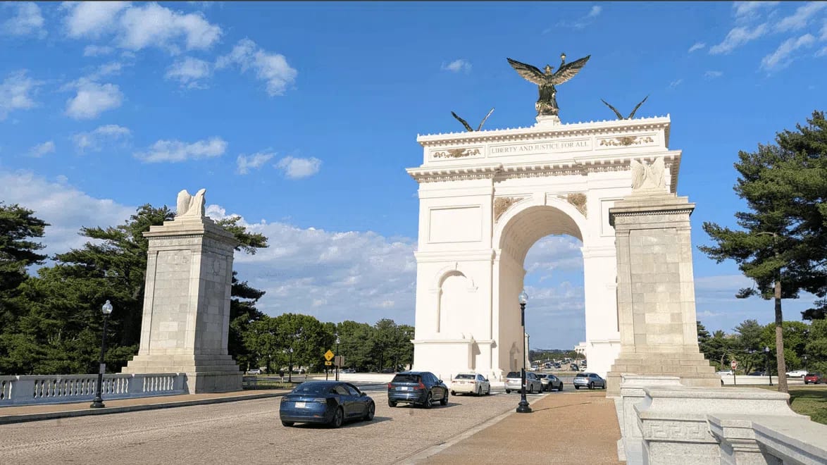 This digital rendering depicts the proposed neoclassical United States Triumphal Arch, an immense white stone monument spanning a multi-lane roadway filled with passing sedans. Positioned along the Arlington Memorial Bridge approach near Memorial Circle, the structure features a large central barrel-vaulted arch flanked by detailed architectural columns and decorative side niches. The top frieze of the main structure is inscribed with the phrase "LIBERTY AND JUSTICE FOR ALL" and is crowned by three prominent golden sculptures, including a large, central winged figure holding a torch and crown aloft, flanked by two smaller winged figures. Separate, heavy stone pylons with sculpted eagle motifs stand on either side of the entrance lane, framing the gateway against a bright blue sky filled with scattered white clouds.