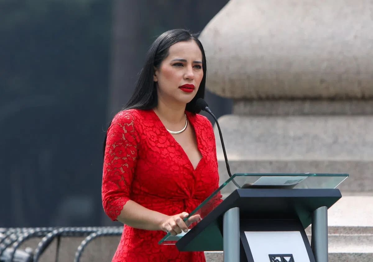 This image features Mexican politician Sandra Cuevas delivering a speech at a podium. She is wearing a striking scarlet red floral lace dress that has three-quarter length sleeves, a deep V-neckline, and a wrap-style bodice. Her look is complemented by bold red lipstick, straight dark hair, and a simple gold necklace. Cuevas stands behind a modern, clear-topped podium with two microphones, positioned in front of a large, light-colored stone structure. 