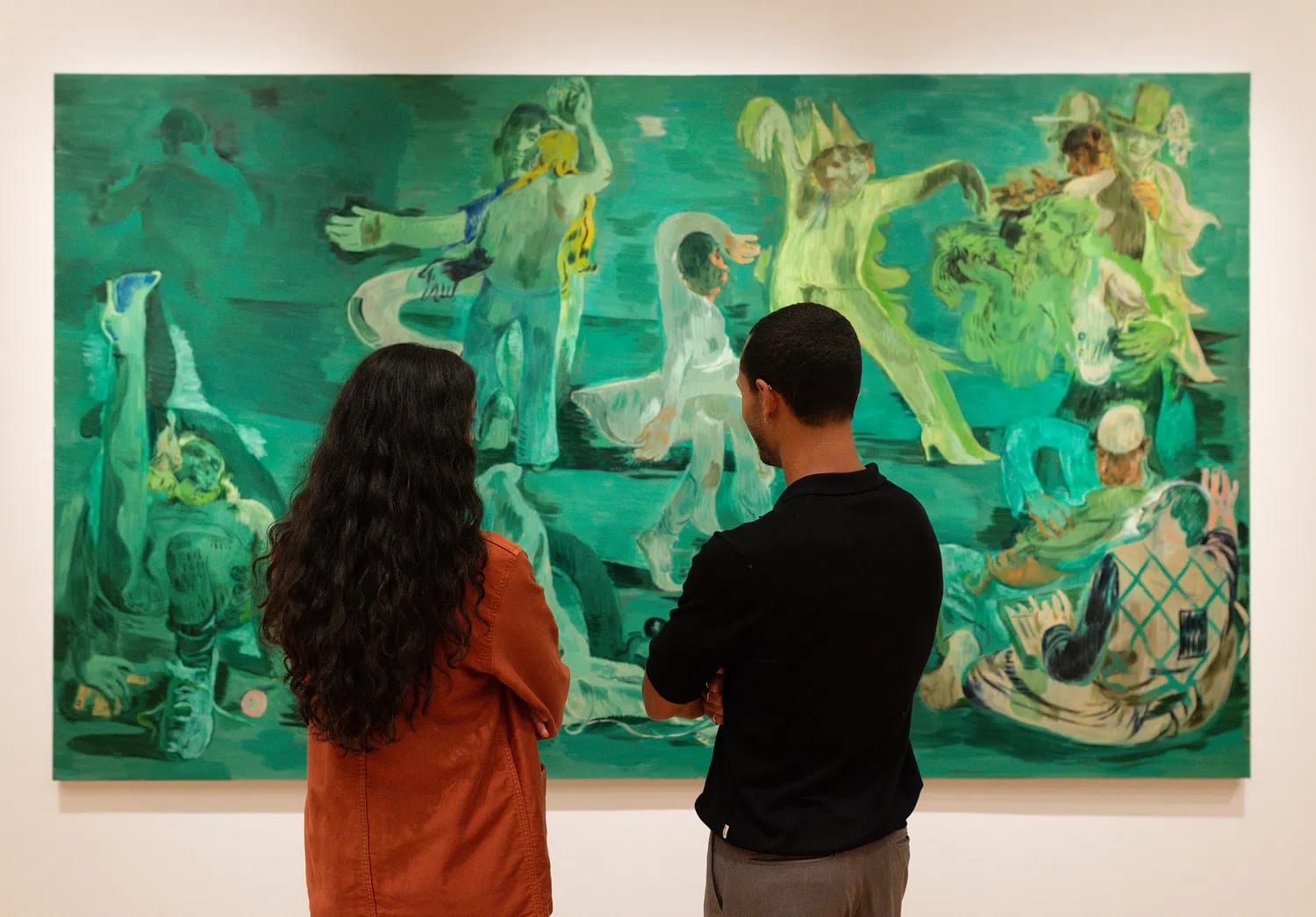 A wide gallery shot captured inside The Courtauld Gallery shows two gallery visitors—a woman with long, dark curly hair wearing an orange jacket on the left and a man in a black long-sleeved shirt on the right—standing with their backs to the camera as they closely study a massive figurative painting. The artwork, titled Ghost Ball (2023) by celebrated artist Salman Toor, dominates the frame with its signature, surreal palette of deep emerald and absinthe greens. Across the vast canvas, a series of loose, fluid, and elongated figures are depicted in various expressive, theatrical poses that suggest a dreamlike nocturnal gathering or party. The figures—some wearing whimsical caps or costumed elements, others lounging or dancing—inhabit a stylized, safe nocturnal environment that evokes themes of community, intimacy, and queer South Asian experiences. The bright gallery lighting illuminates the clean white wall surrounding the massive canvas, casting a subtle warmth on the shoulders of the spectators engrossed in the scene.