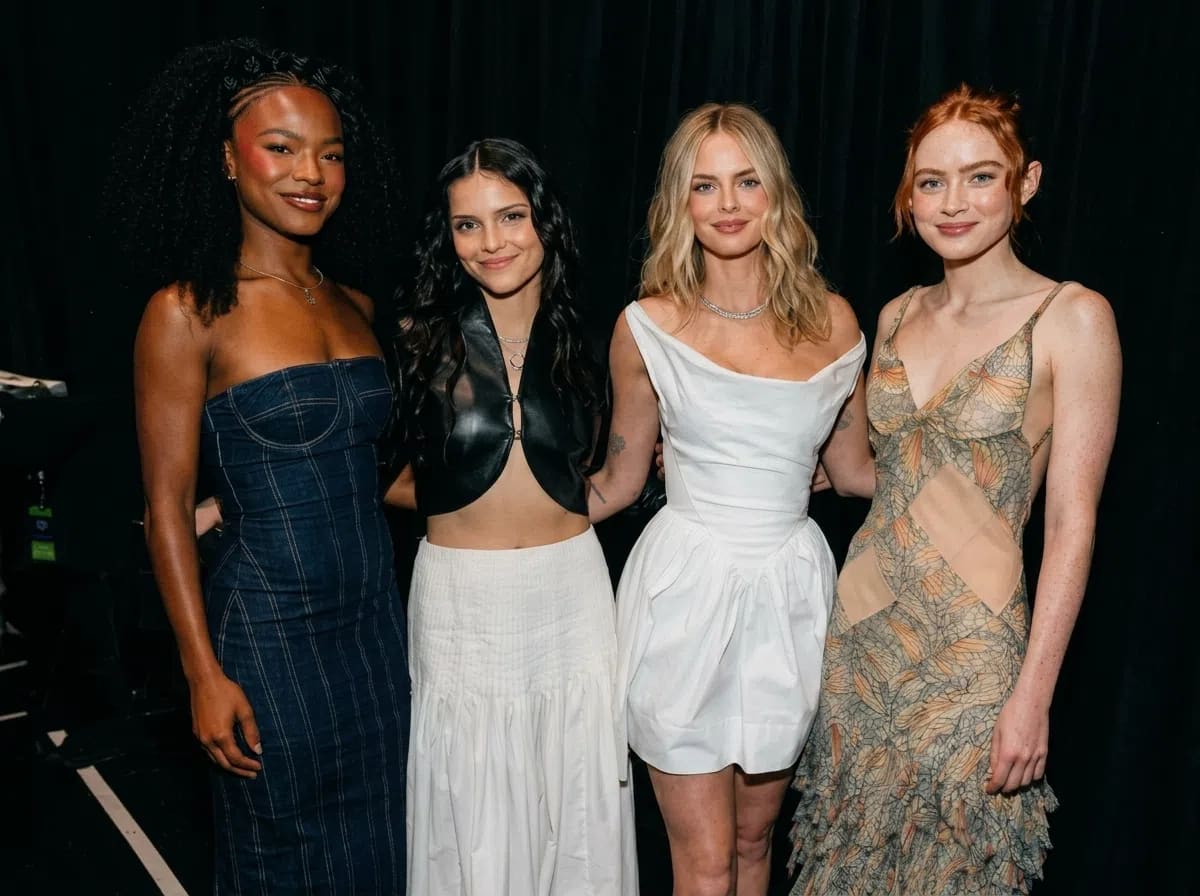A medium-shot portrait captures Maya Boyd, Inde Navarrette, Samara Weaving, and Sadie Sink standing closely together against a dark, plain black backdrop. On the far left, Maya Boyd poses with a soft smile, styled with voluminous, dark curly hair pinned back at the sides. She wears a dark blue strapless denim maxi dress featuring visible pinstripes and structured seam lines along the bodice. Standing next to her, Inde Navarrette smiles directly at the camera with long, wavy dark hair framing her face. She wears an edgy black leather or faux-leather halter-neck crop top that splits open at the lower front, paired with a high-waisted, flowing white maxi skirt. Next in line, Samara Weaving smiles subtly, showcasing shoulder-length blond waves parted down the middle. She wears a structured, sleeveless white cocktail dress with a draped cowl neckline, a fitted waist, and a slight balloon silhouette at the skirt, complemented by a delicate silver necklace. On the far right, Sadie Sink smiles warmly with her signature red hair styled into an elegant updos with loose strands framing her face. She is dressed in an intricate, sleeveless V-neck maxi dress adorned with semi-sheer mesh paneling across the midriff and tiered, ruffled fabric detailing in muted tones of beige, green, and orange. The lighting is direct and bright, casting soft shadows behind the women and highlighting the varying textures of their outfits.